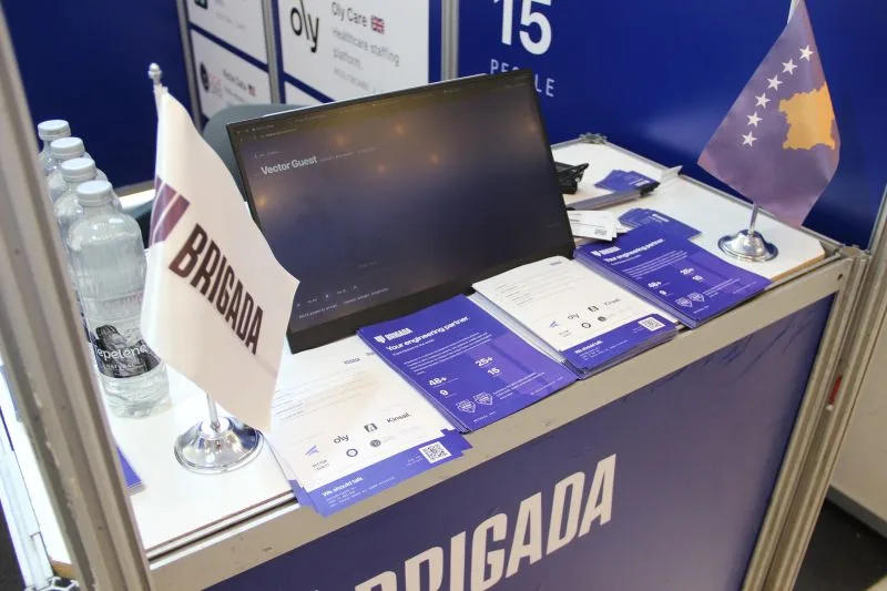 The Brigada stand counter with a laptop, Brigada and Kosovo flags, and printed case study sheets.