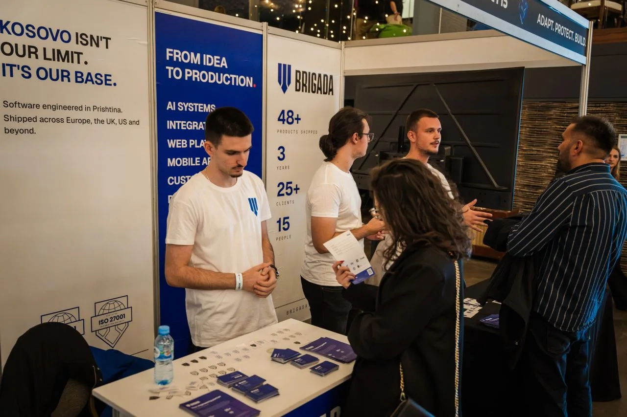 Two of the Brigada team at the booth counter, talking with visitors beside a banner listing AI systems, web platforms and mobile apps.
