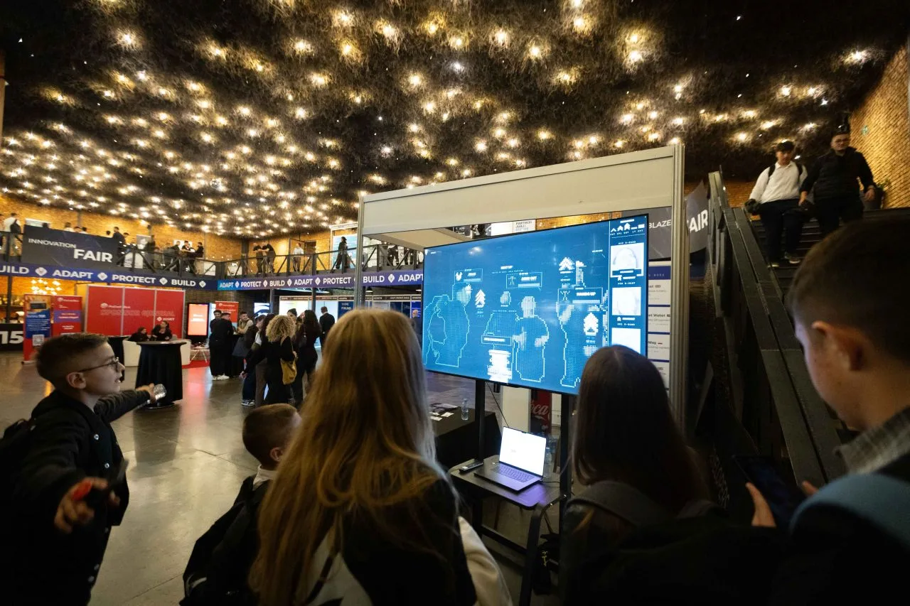 Visitors playing the Brigada game on the booth screen, the arena's lights overhead.