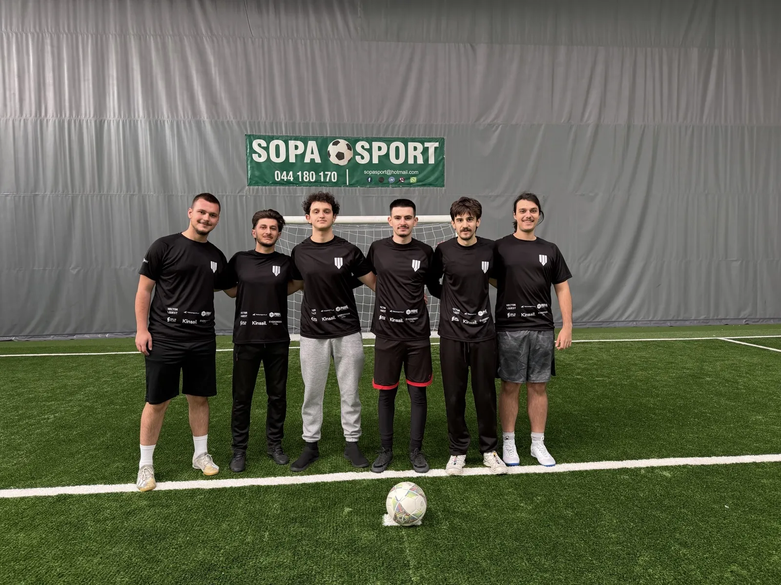Six members of the Brigada team in black Brigada shirts, arm in arm in front of a goal, a ball on the line in front of them.